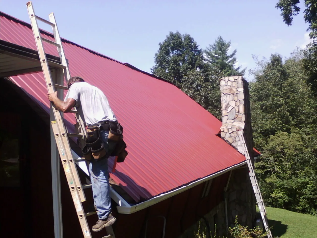 Roofer on a ladder beside a red metal roof and stone chimney — Soffit & Fascia Repair by Roofing Brothers in Gibbstown